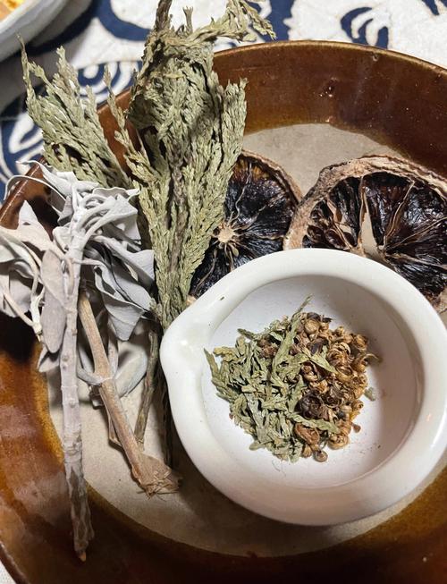A rustic ceramic bowl containing dried herbs, including sage, cedar, and seed pods, along with slices of dried citrus and a small white mortar holding a herbal blend, used for incense or traditional cleansing rituals.