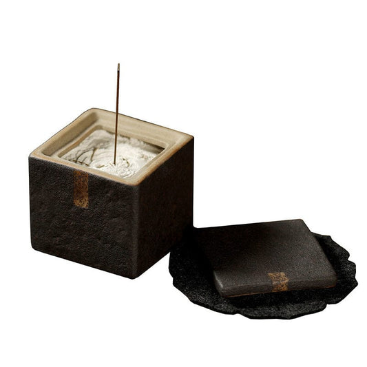 A square incense holder with matte ceramic texture and brass accents, resting on a wooden surface with incense smoke rising gently—embodying stability, wabi-sabi, and Earth Element presence.