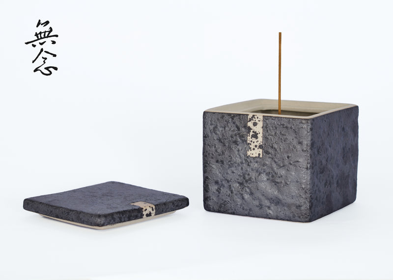 Grey cube-shaped incense burner with an upright stick and lid beside it.