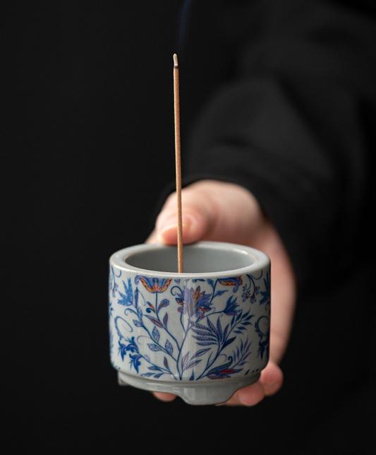 Hand holding a blue and white floral holder with a lit incense stick.