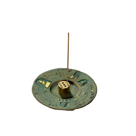 A round ceramic incense tray carved with Buddha figures, finished in weathered turquoise with a central brass socket—evoking ancient stillness, sacred protection, and elemental harmony.