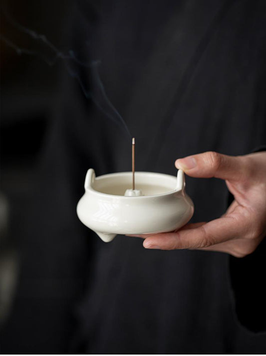 Hand holding a minimalist white ceramic Chinese incense burner with a lit natural incense stick, releasing gentle smoke — symbolizing mindfulness and Zen tranquility.