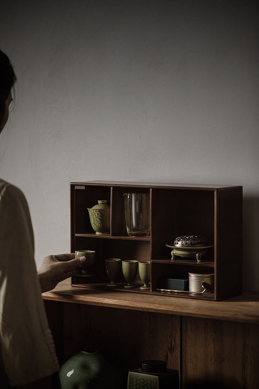 Person arranging ceramic tea cups on a wooden display shelf with incense burners and glassware in a minimalist, Zen-inspired setting.