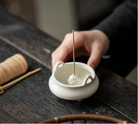 A hand placing a burning incense stick into a white ceramic incense holder on a dark wooden table, creating a calm and Zen-inspired atmosphere.