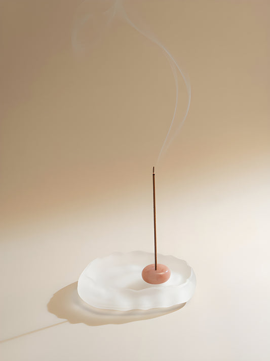 A slender rose incense stick burning in a pink round holder on a white dish in warm sunlight.