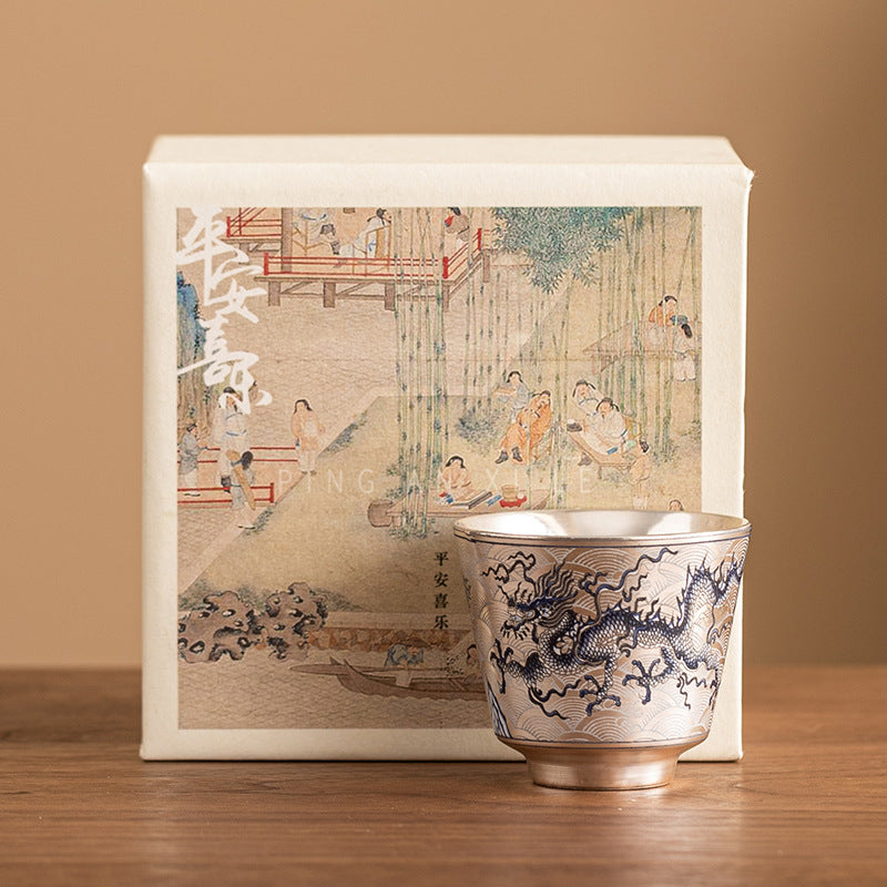 Ceramic cup with dragon design in front of a decorative box with traditional artwork on a wooden surface.