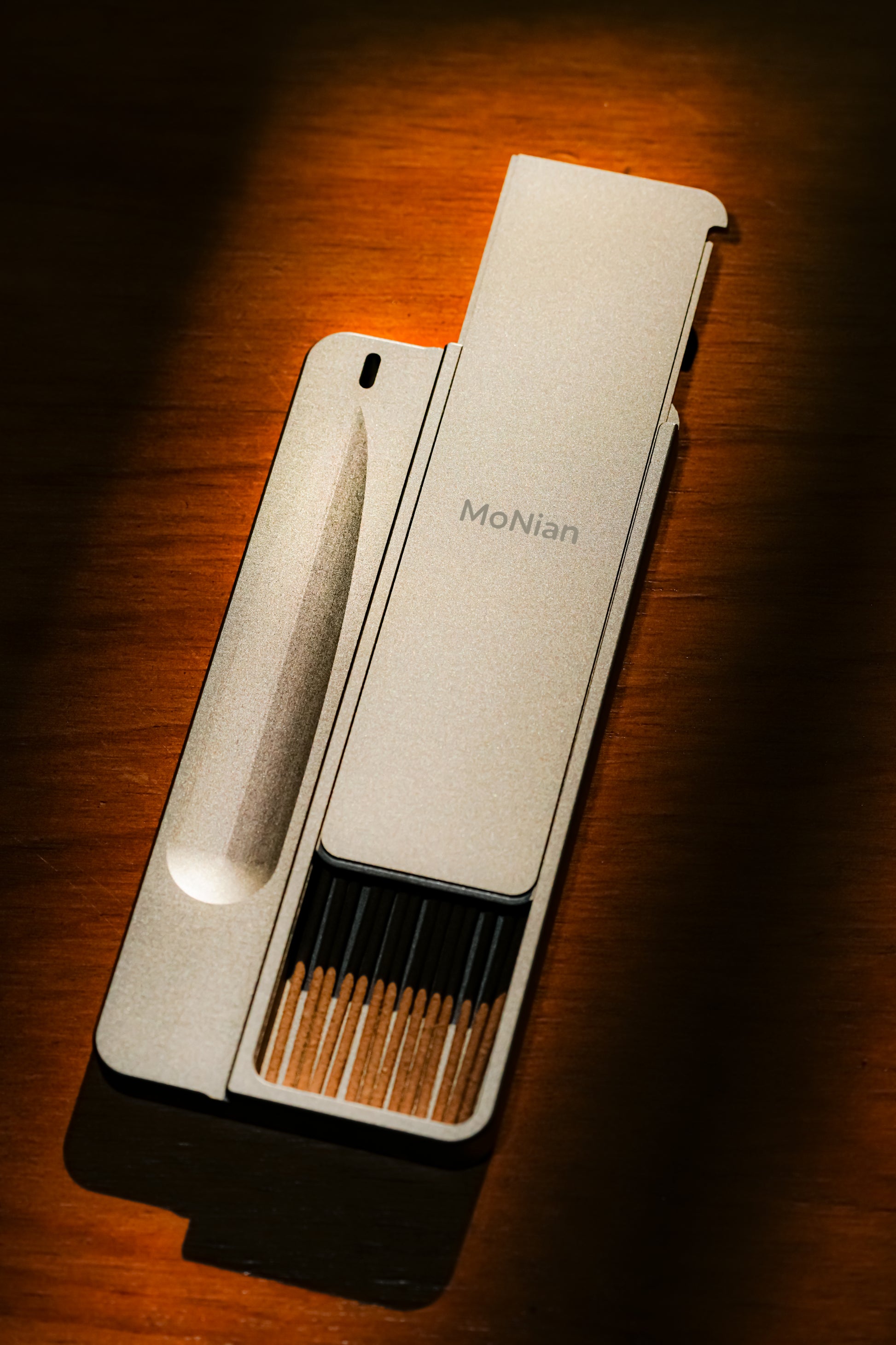 Sleek rectangular metal incense burner on a wooden table with a single incense stick angled upward in a recessed groove, faint smoke rising.